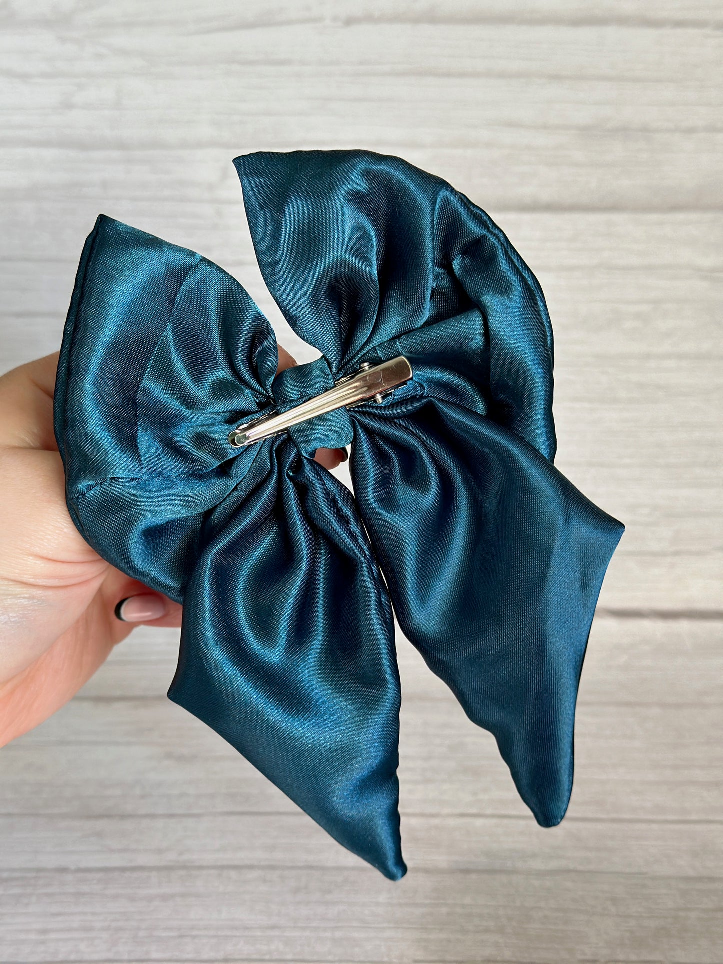 A hand holding the Crafts by Kate "Silk Sailor Bow Clip" in Teal Blue, featuring a crocodile clip on the back, is showcased against a light wooden background.