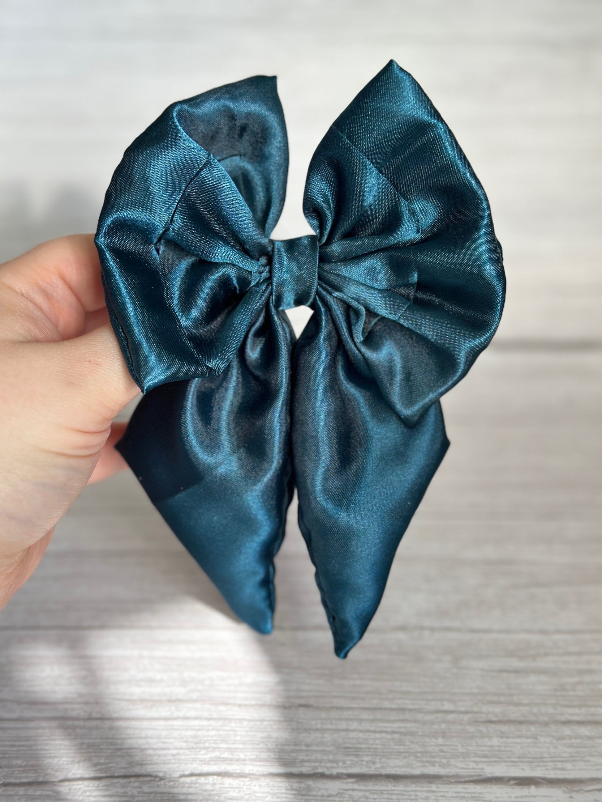 A hand holds the Crafts by Kate Silk Sailor Bow Clip in teal blue against a light wooden background. The bow, secured with a crocodile clip, showcases two loops and long tails, with a slightly crumpled texture.