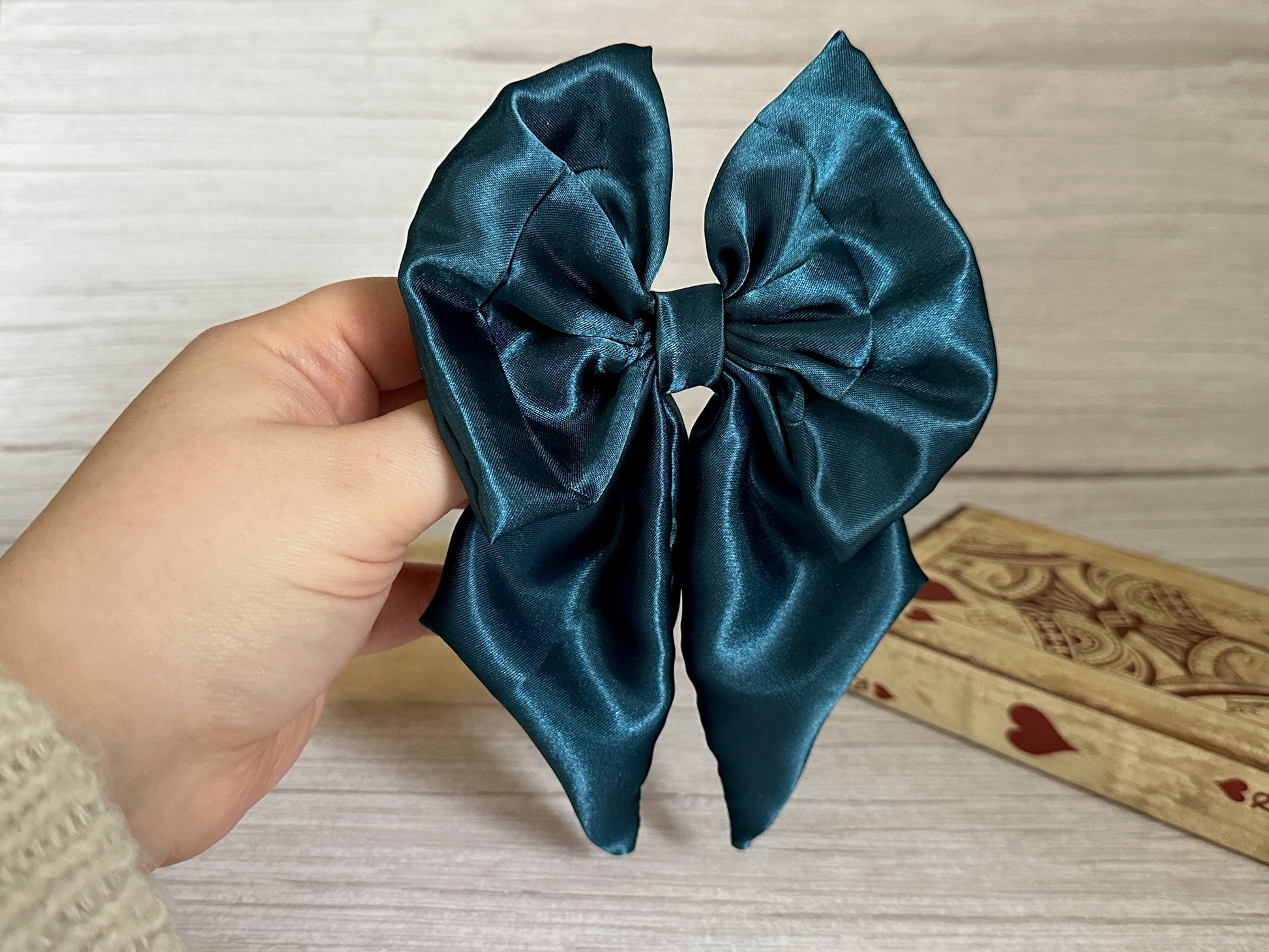 A hand is holding the large, glossy Silk Sailor Bow Clip in Teal Blue from Crafts by Kate against a light wood background, with a wooden box featuring heart designs partially visible on the right.