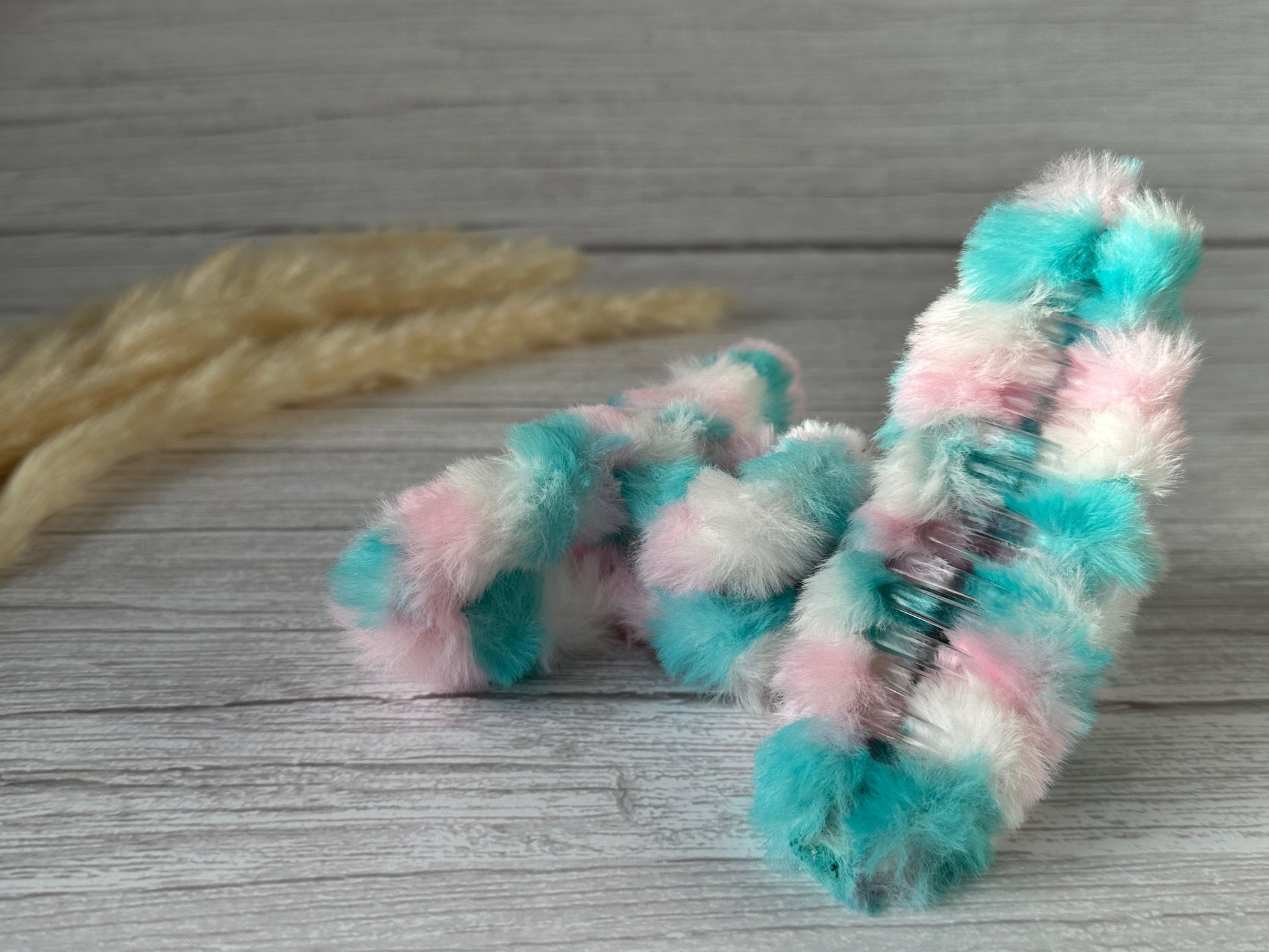 The Fluffy Hair Claw Clip - Unicorn Dreams by Crafts by Kate, featuring a pastel color scheme of pink, blue, and white, sits on a wooden surface. The background includes beige pampas grass, adding a natural element to the scene.