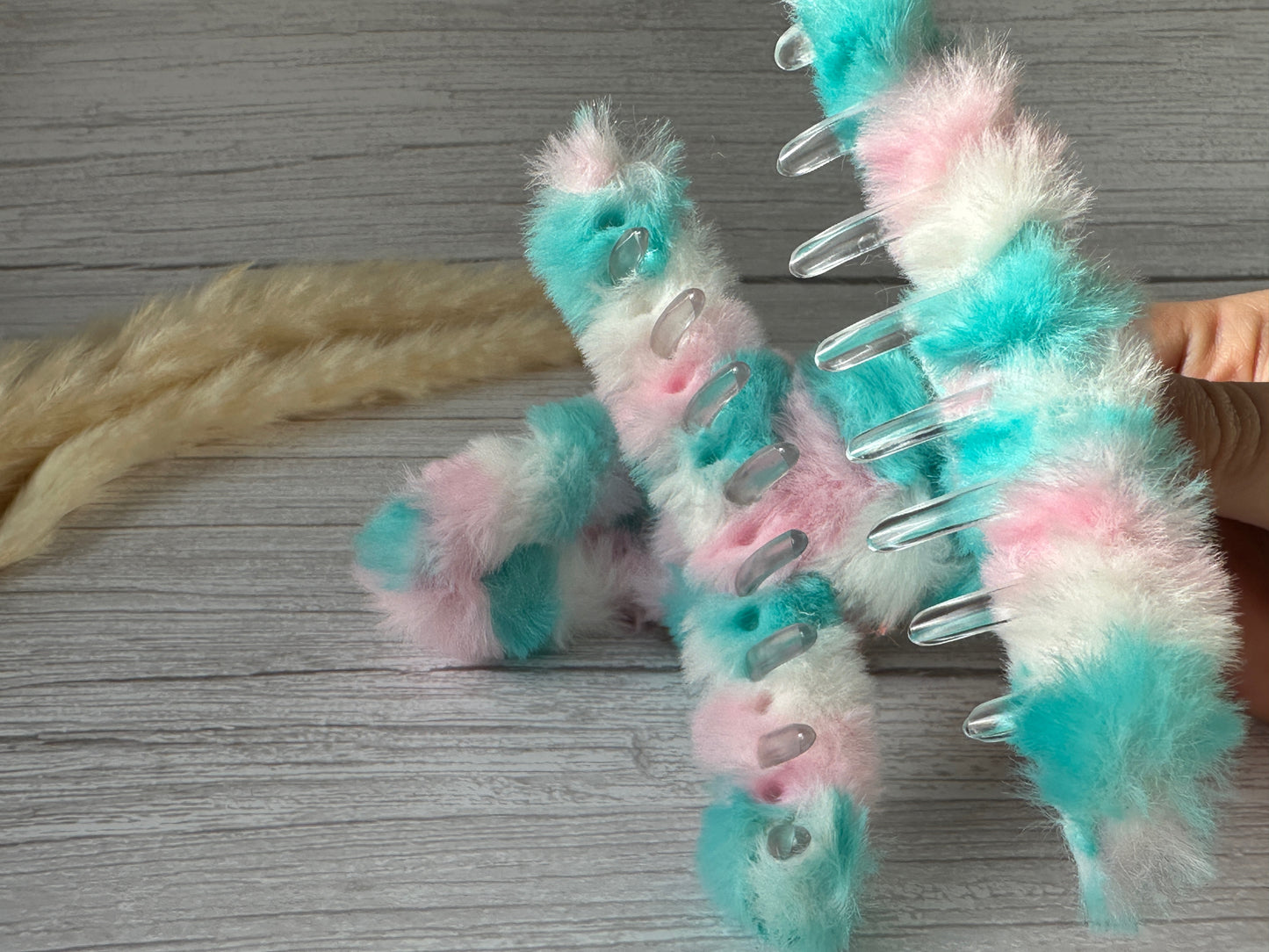 A hand holding a fuzzy, multicolored handcrafted hair claw clip called "Unicorn Dreams" by Crafts by Kate, which features teal, pink, and white segments and transparent prongs. A second similar clip and a luxurious cream-colored plush fabric object are placed on a light wooden surface in the background.