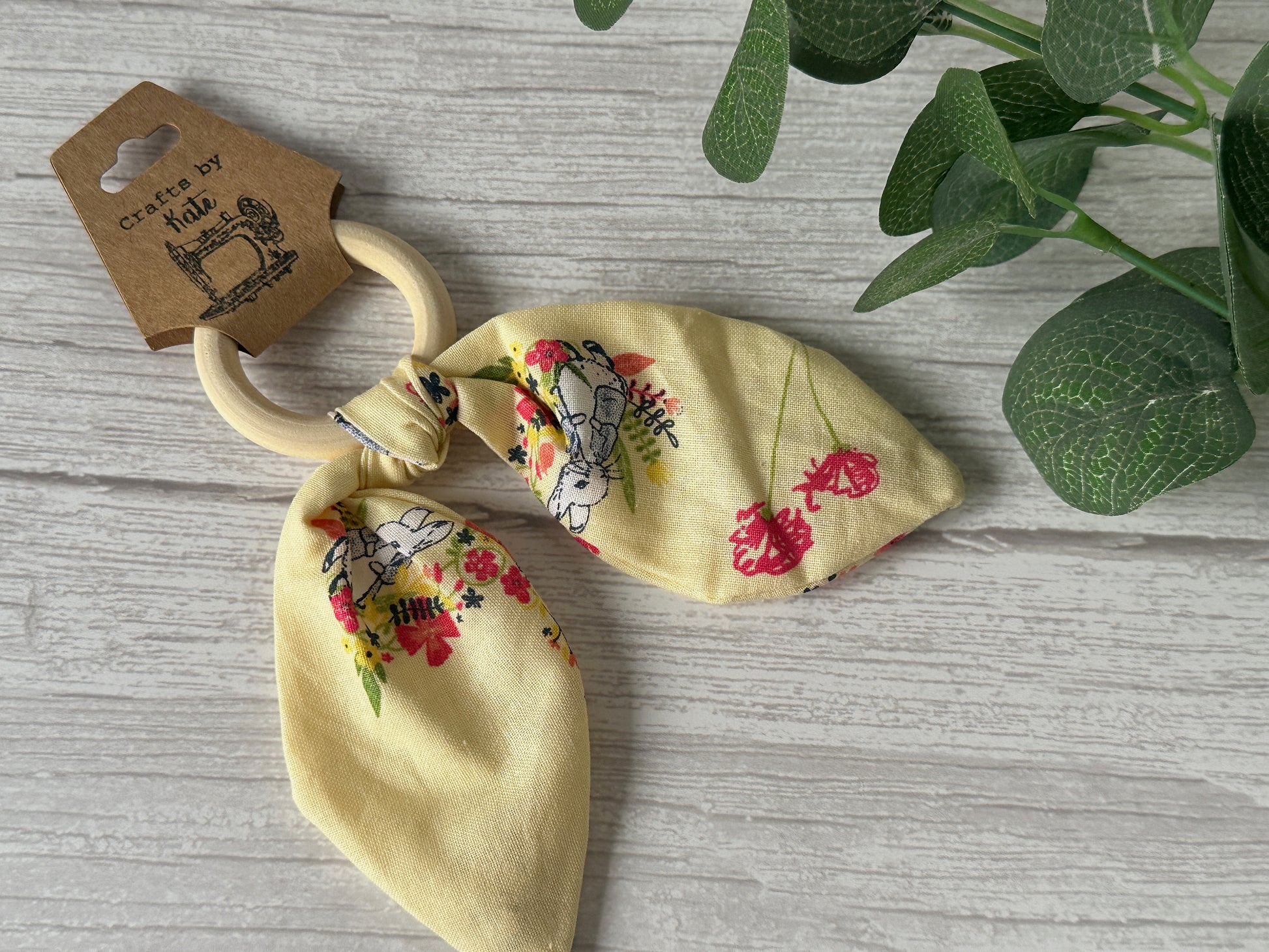 A Cotton & Bamboo Baby Teething Rattle, featuring a lemon floral fabric with Peter Rabbit illustrations from Crafts by Kate, lies on a light wooden surface next to a green potted plant. The brown tag attached to the rattle highlights the brand name "Crafts by Kate.