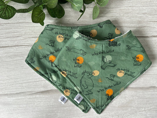 Two Cotton & Terry Towelling Baby Bandana Bibs in green, featuring woodland-themed illustrations and text from Crafts by Kate, displayed on a light wood surface. The handcrafted baby bibs showcase sketches of animals like a fox, snake, and birds with phrases such as "Oh help! Oh no! It's a gruffalo!" and "Why, Fox, hello!" A leafy plant is above the bibs.