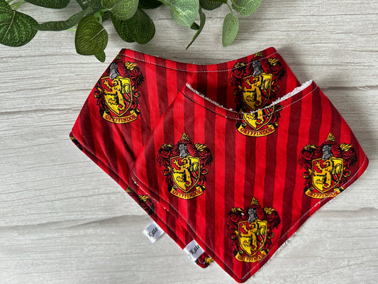 The image features two bandana bibs from the Harry Potter Houses Bandana Bib Set by Crafts by Kate. Each red and gold striped cloth bib prominently displays the Gryffindor house crest, which includes a lion insignia from the Harry Potter series. Green leaves can be seen in the background.