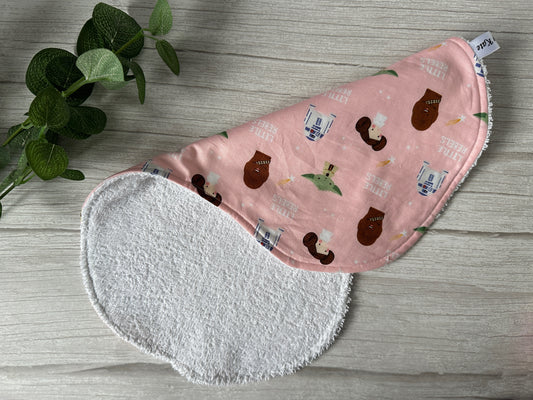 A baby burping cloth made from ultra-soft cotton, featuring a pink design adorned with charming Star Wars Little Rebels characters and phrases. The item, designed by Crafts by Kate, includes a soft white terry cloth backing and is showcased on a light wooden surface beside a small, leafy green plant.